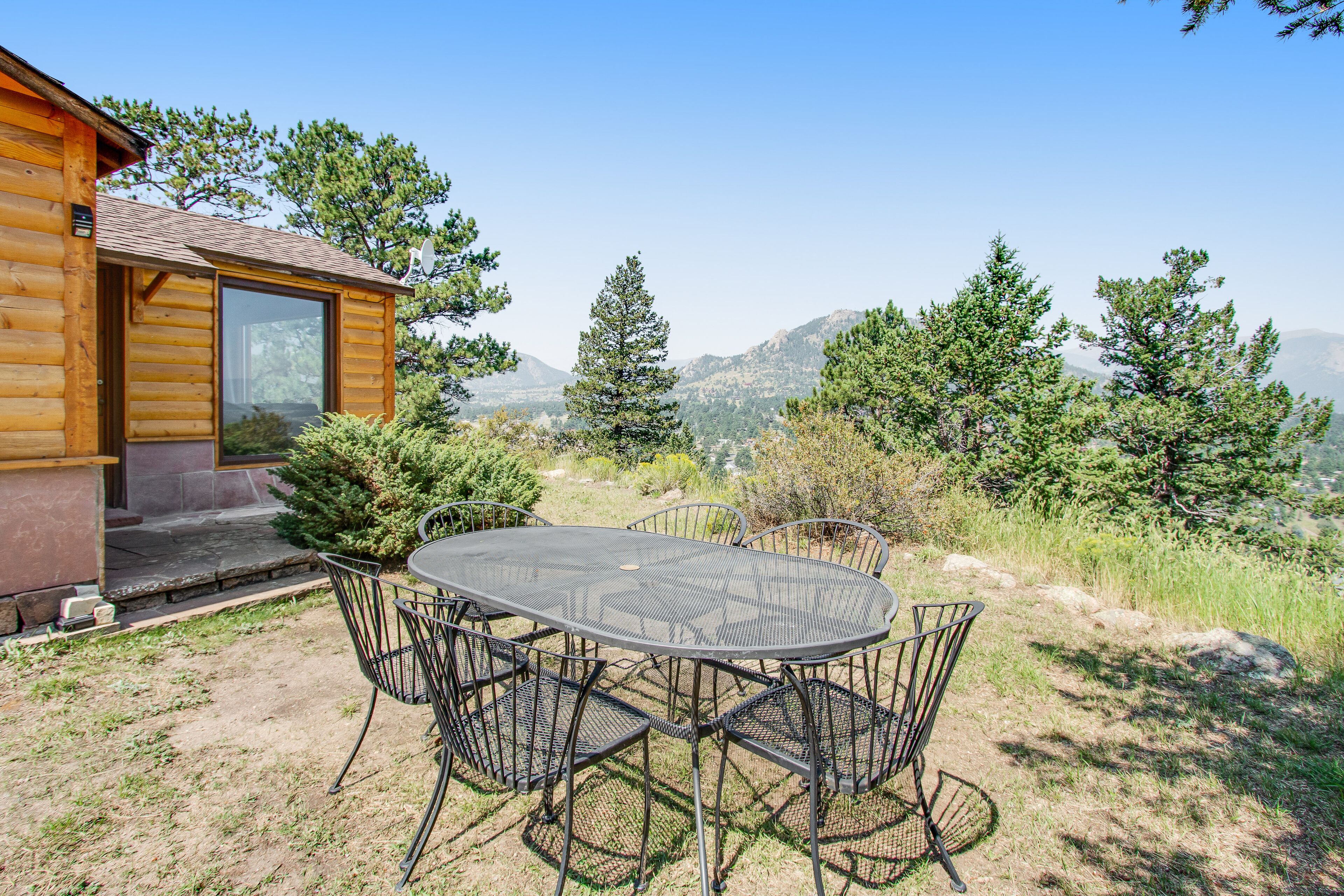 A wooden cabin with a patio table and chairs, surrounded by trees and mountains.