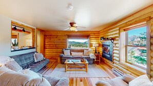 A log cabin interior with a sofa, armchairs, a coffee table, a fireplace, and large windows offering a view of the outdoors. - Memorable mountain condo with 2 fireplaces, lovely views, W/D, & pool access (Estes Park)