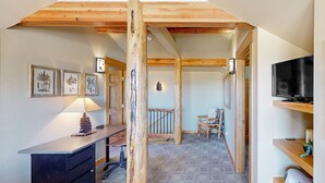 A cozy room with a desk, a lamp, framed botanical prints, a TV, and wooden beams. - Memorable mountain condo with 2 fireplaces, lovely views, W/D, & pool access (Estes Park)