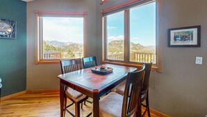 A dining area with a wooden table and chairs, large windows with a view of mountains, and a clock on the wall. - Two BR condo w/ Brand NEW HOT TUB and great views! Community pool perfect for kids (Estes Park)
