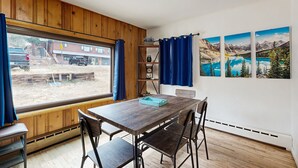 Et spiseområde med et træbord og stole, udsigt udad samt tre landskabsmalerier på væggen.