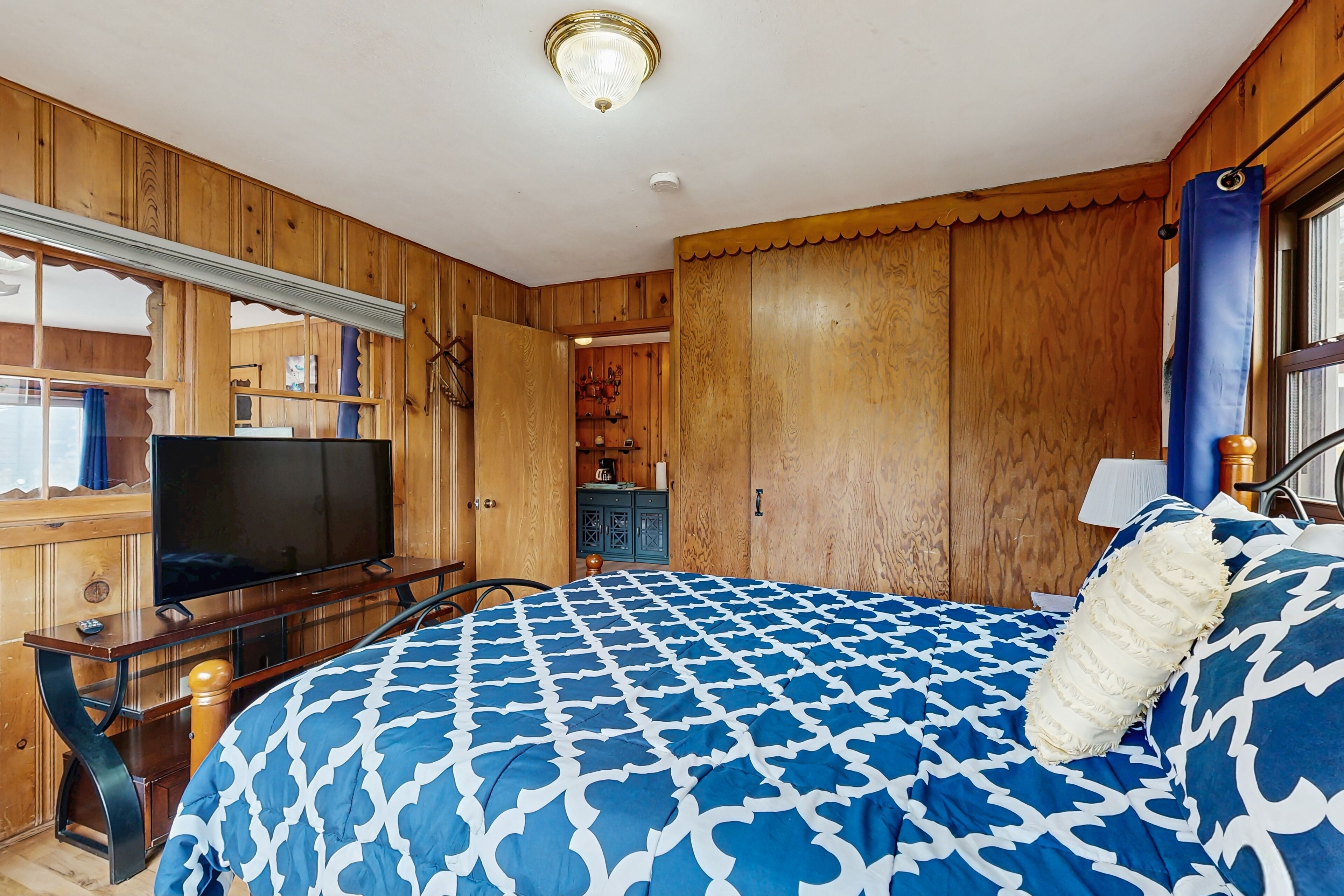A bedroom with wooden paneling, a bed with a blue and white patterned bedspread, a television on a wooden stand, and a window with a blue curtain.