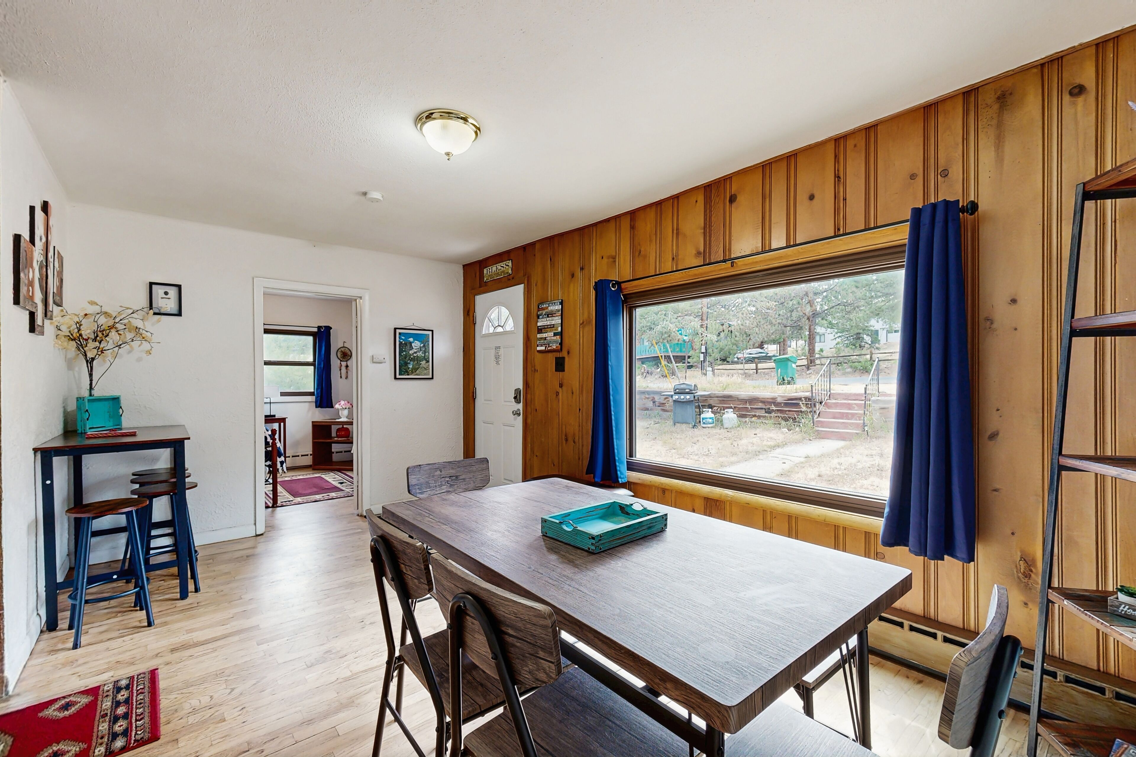 Un coin repas avec une table et des chaises en bois, une fenêtre ornée de rideaux bleus et une vue sur l’extérieur.