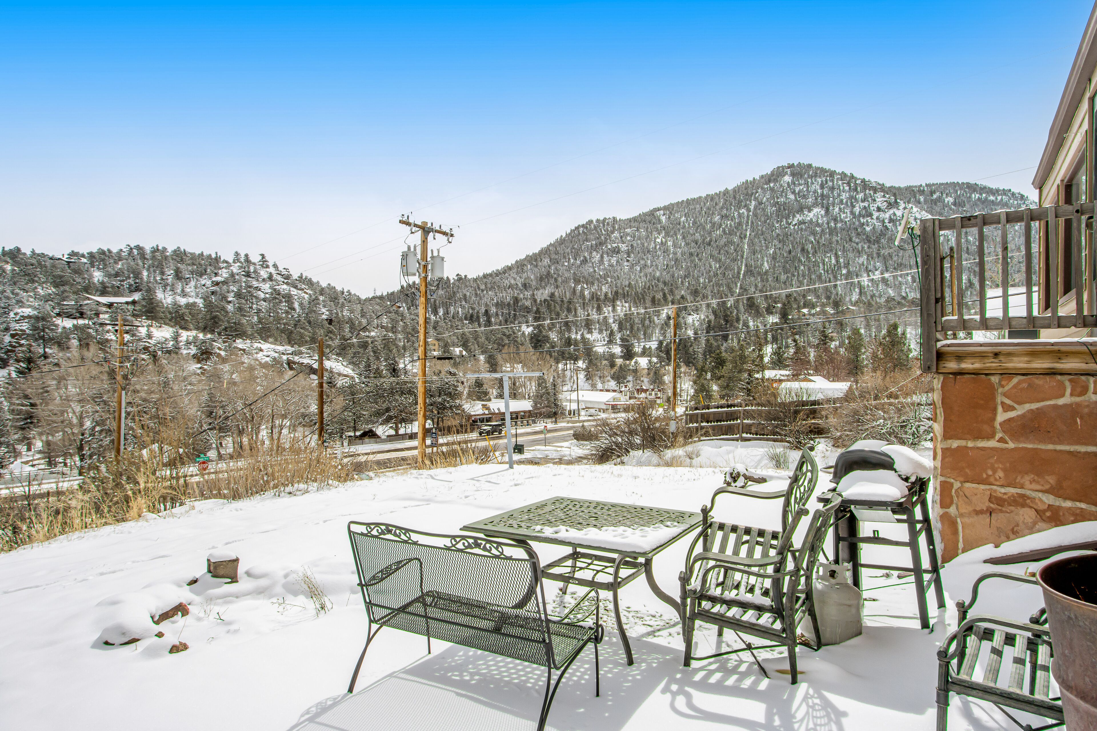 A snow-covered patio with metal furniture, a stone wall, and a view of a snowy mountain.