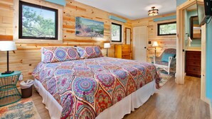 A wooden cabin bedroom with a large bed, two windows, a painting, and a small table with a lamp. - Charming cabin with full kitchen, furnished deck, & incredible views (Estes Park)