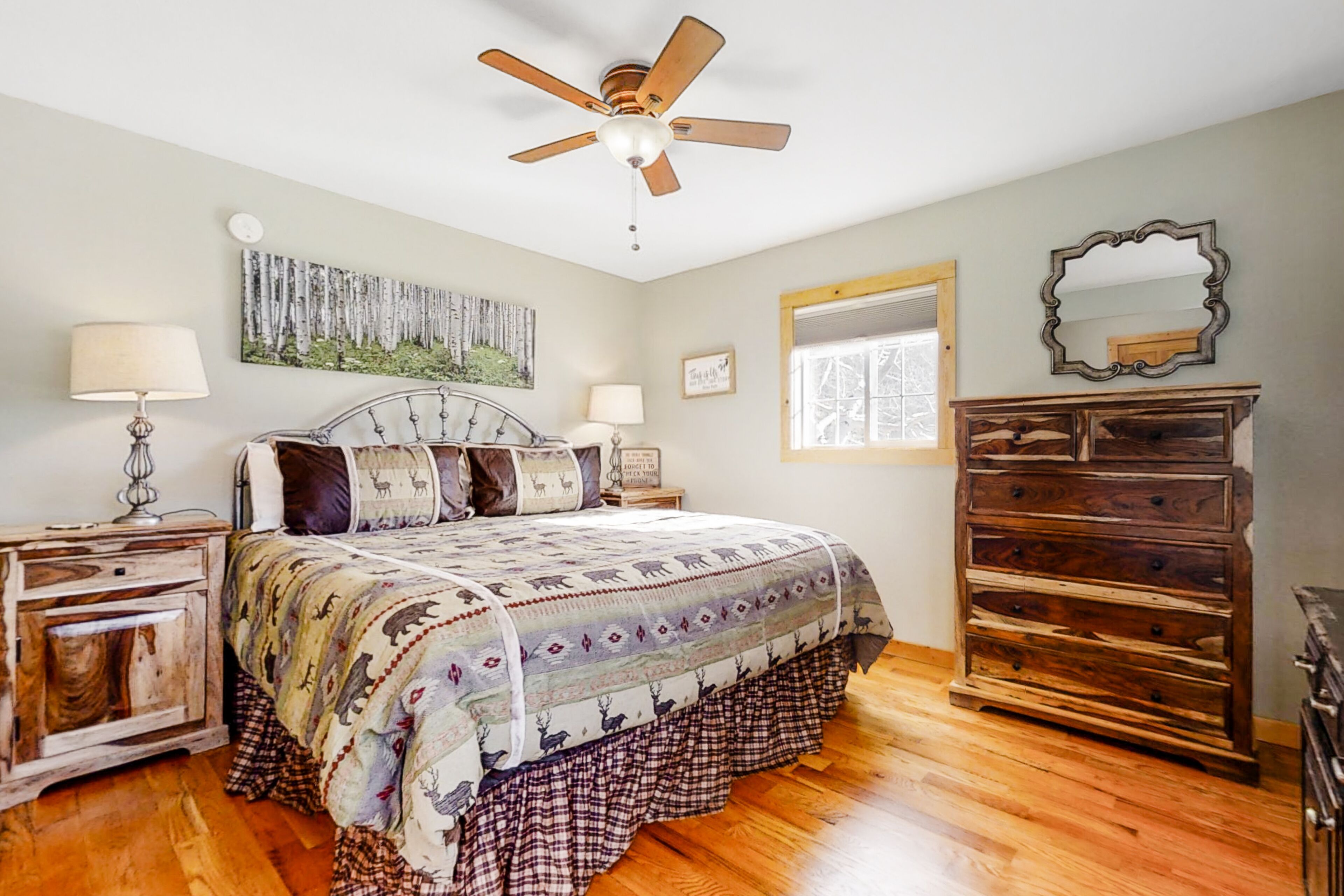 A bedroom with a wooden floor, a bed with a patterned bedspread, a ceiling fan, a dresser, a mirror, and a window with a view of trees.