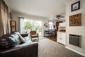 A living room with a leather couch, a dining area with a table and chairs, a kitchen with cabinets and a ceiling fan, and a large window with curtains. - Hilltop Haven at Sommer Dorf (West Branch)