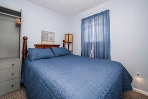 A bedroom with a bed, a dresser, a nightstand, a lamp, and a window with curtains. - Hilltop Haven at Sommer Dorf (West Branch)