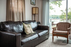 Ein dunkelbraunes Ledersofa mit einem einzelnen Zierkissen, ein Holzstuhl und Blick auf ein Gewässer durch ein Fenster.