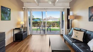 A living room with a sofa, a coffee table, and a dining table. There is a sliding glass door leading to an outdoor patio with a view of a house and greenery. - KBS A107 - Updated Studio next to Beach (Kihei)