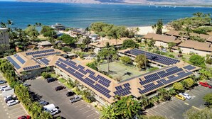 A building with solar panels on the roof, surrounded by parking and greenery, with a view of the ocean and mountains in the background. - KBS A107 - Updated Studio next to Beach (Kihei)