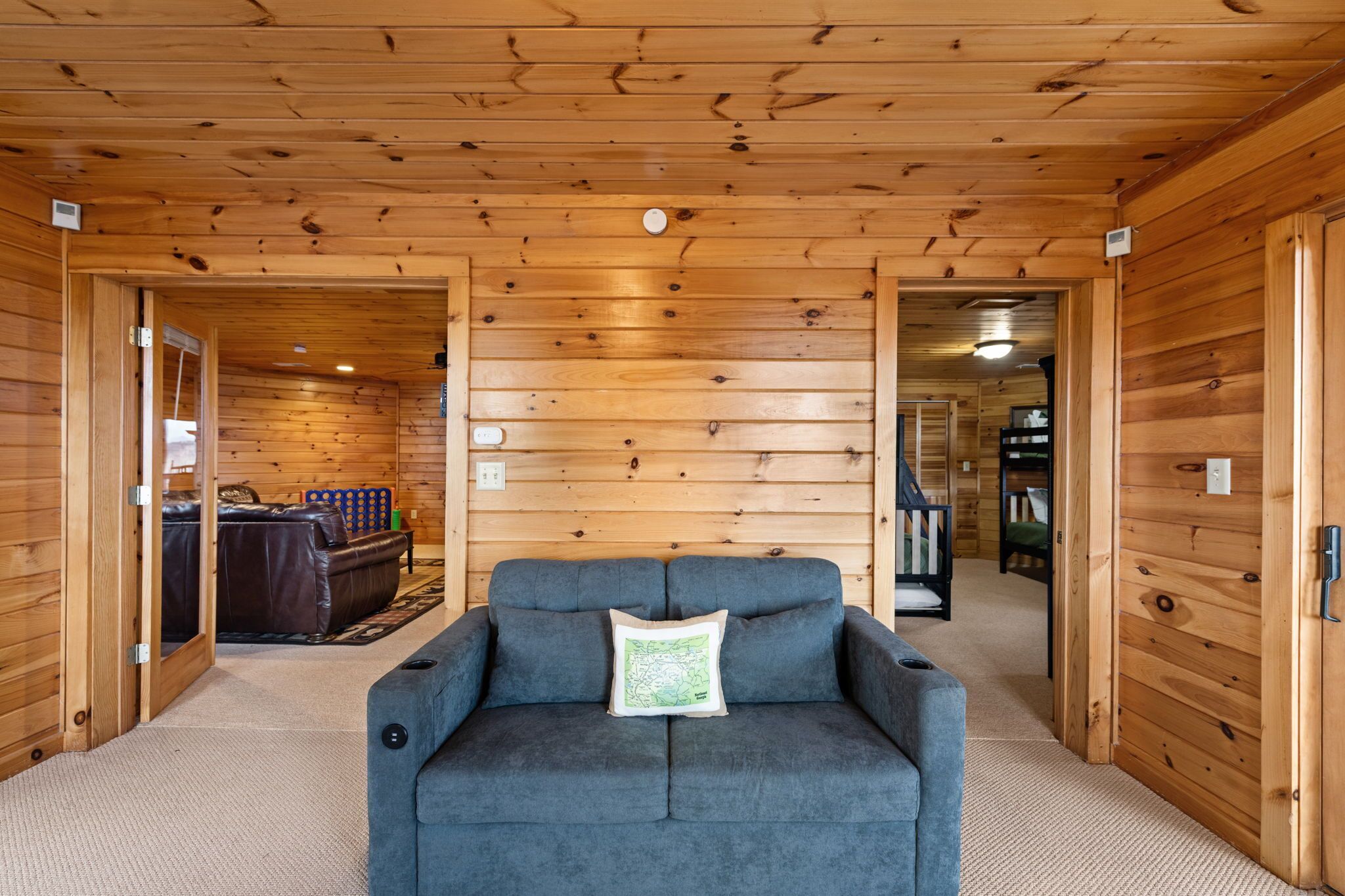 Habitación con paneles de madera, un sofá gris y un cojín con un mapa, una puerta de madera, y un pasillo visible que conduce a otra habitación.