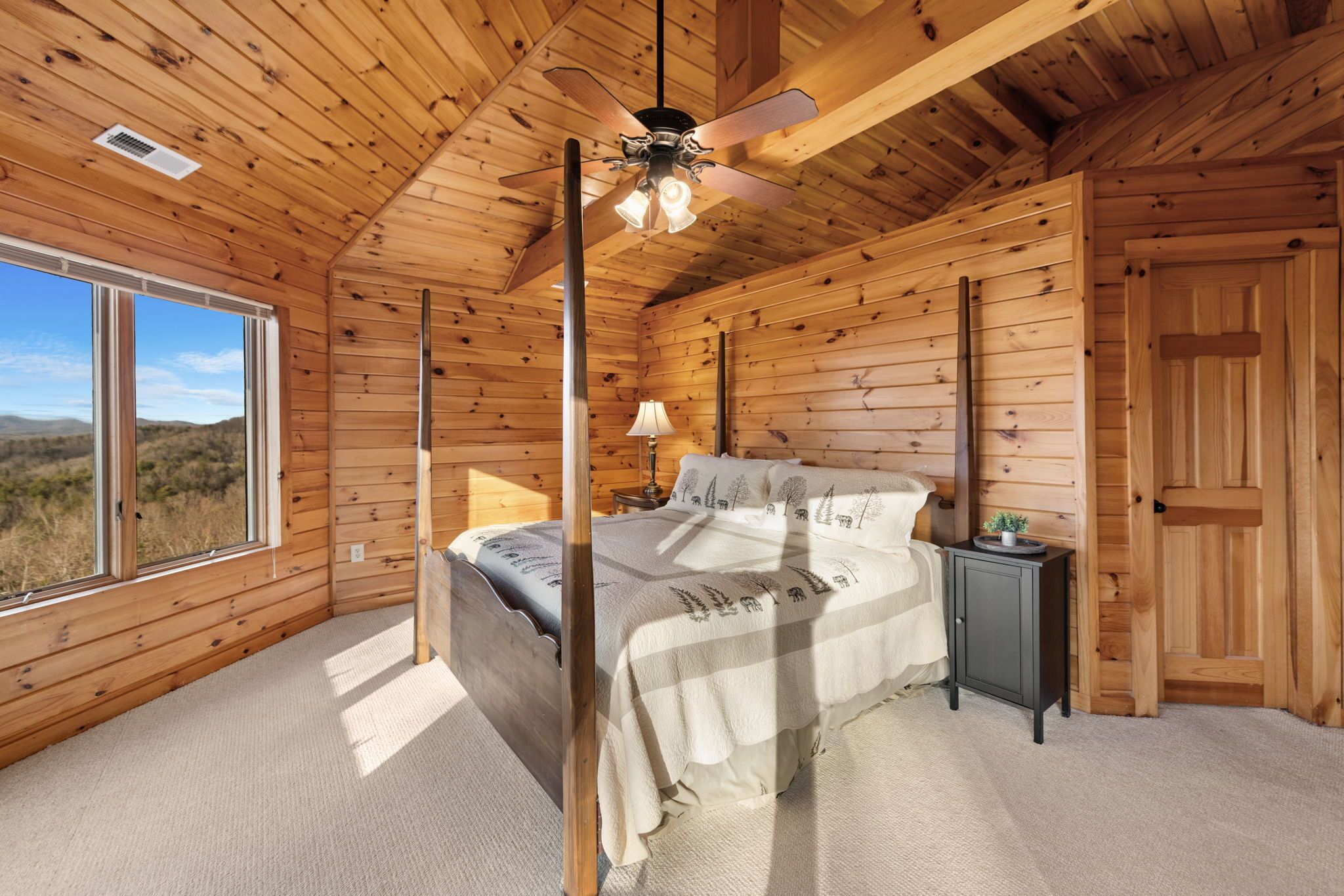 Een slaapkamer in een houten chalet met een bed, een nachtkastje, een plafondventilator en een raam met uitzicht op de natuur.