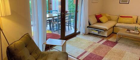 A living room with a sofa, a small table, a chair, and a view through a glass door to an outdoor dining area.
