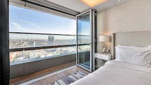 Une chambre d’hôtel moderne dotée d’une grande fenêtre donnant sur la ville, d’un lit avec du linge de lit blanc, d’une table de chevet avec une lampe et d’une porte en verre qui mène à un balcon extérieur.