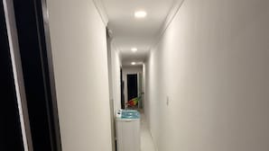 Un couloir étroit et bien éclairé, avec des murs blancs et un sol carrelé. On aperçoit une machine à laver au loin, et une porte est entrouverte sur la gauche.