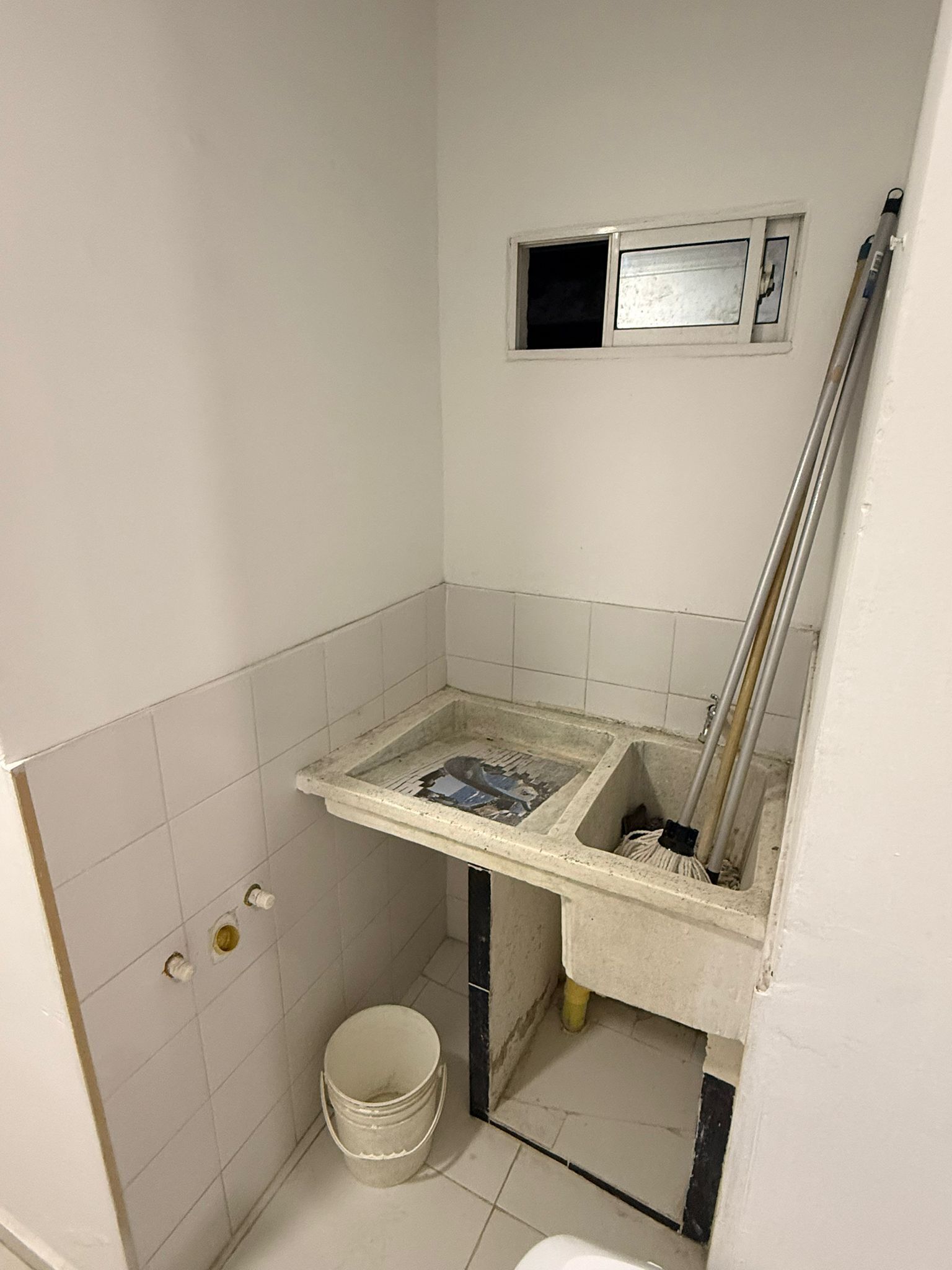 Un baño pequeño con un lavamanos, un balde y una escoba apoyada en la pared.