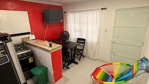 Une cuisine compacte équipée d’une cuisinière à gaz, d’une petite poubelle et d’un coin repas avec une table et des chaises. Un fauteuil à bascule coloré est placé dans l’espace salon.