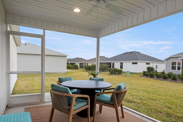 A covered patio with a round table and wicker chairs, overlooking a grassy yard with houses in the background.
