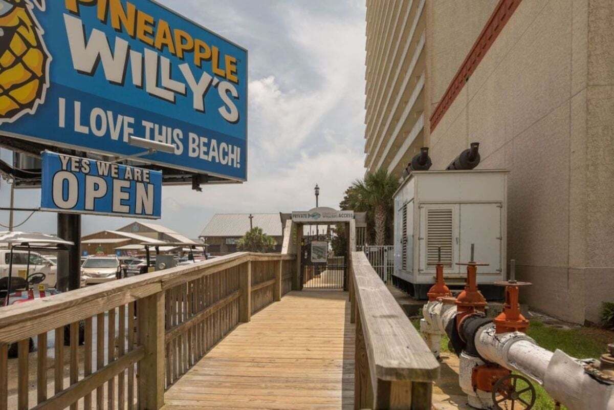 Een houten looppad dat naar een strand leidt, met een bordje voor 'Pineapple Willy's' en een bord waarop staat 'open'.