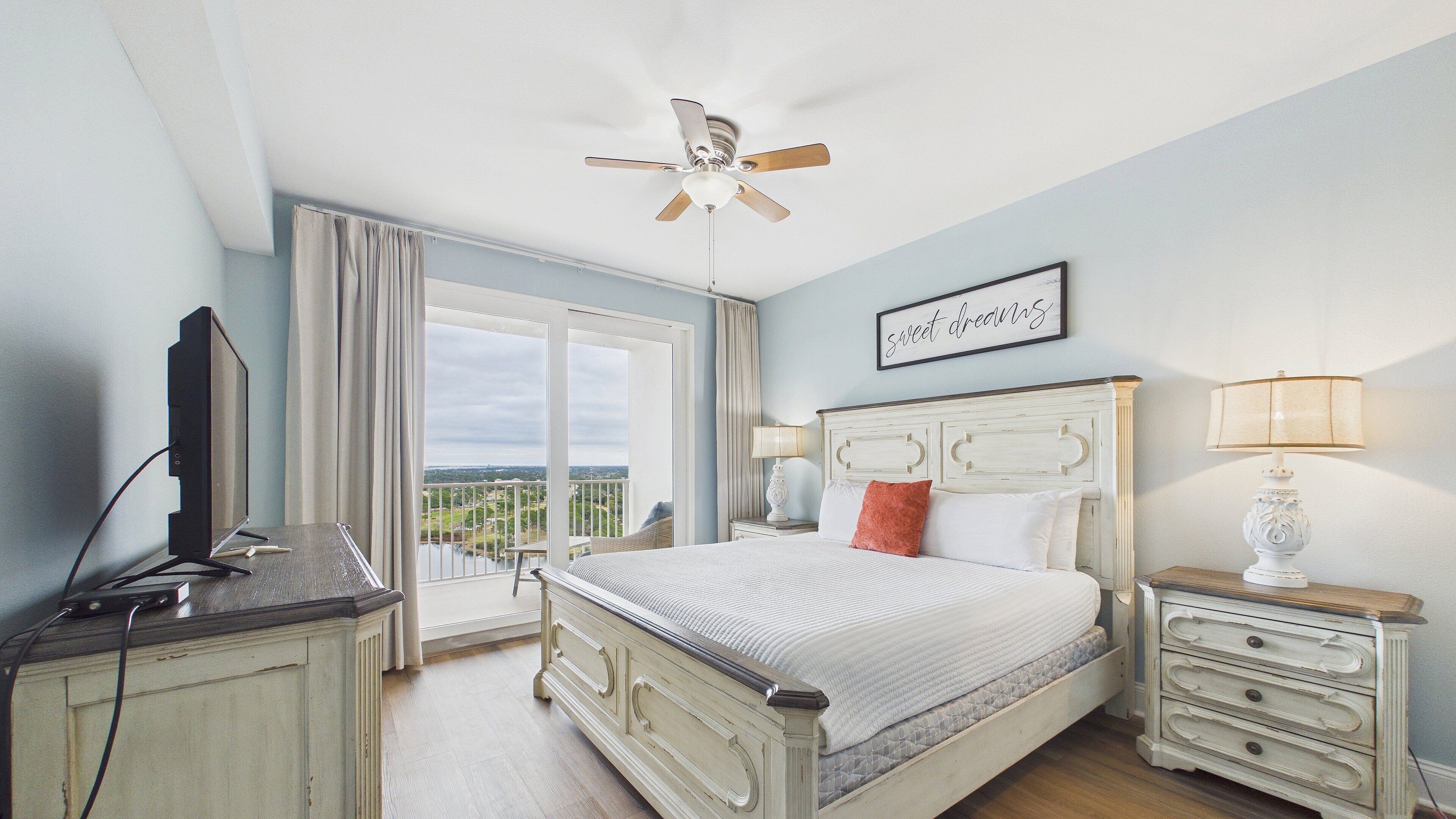 A bedroom with a bed, a nightstand, a lamp, a ceiling fan, and a view of the outdoors.