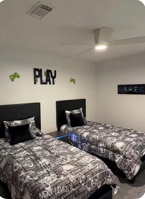 A bedroom with two beds, a ceiling fan, and wall decorations including 'PLAY' and 'GAMER' signs.