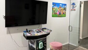 A room with a flat-screen TV mounted on the wall, a gaming console, a pink stool, and a poster of Super Mario characters. - Luxurious, peaceful, & fun 4-bedroom house with a game room in charming La Porte (La Porte)
