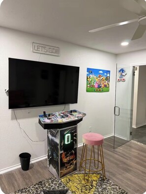 A room with a flat-screen TV mounted on the wall, a gaming console, a pink stool, and a poster of Super Mario characters. - Luxurious, peaceful, & fun 4-bedroom house with a game room in charming La Porte (La Porte)