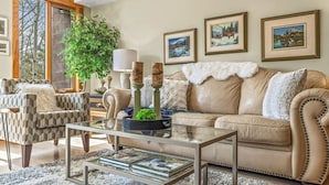 A living room with a beige sofa, patterned armchair, and a glass coffee table with books. There are framed pictures on the wall and a large plant in the corner. - Rustic Creekside Retreat 2BR and 2BT with Stunning View! (Canmore)