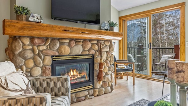A stone fireplace with a TV mounted above it, a cozy seating area with a fireplace, and a large sliding glass door leading to an outdoor deck. - Rustic Creekside Retreat 2BR and 2BT with Stunning View! (Canmore)