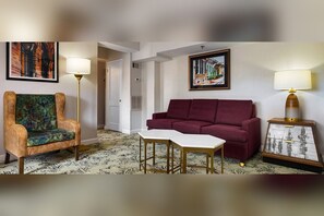 Une pièce avec un tapis à motifs, un canapé rouge, un fauteuil à motifs, une petite table blanche et un meuble avec des tiroirs.