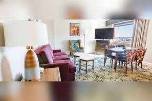 Une chambre d’hôtel avec un canapé, une table à manger avec des chaises, une télévision et une grande fenêtre avec des rideaux.