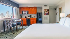 Une chambre d’hôtel avec un grand lit, un bureau avec une chaise, une kitchenette et une vue sur les toits de la ville.