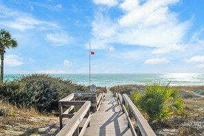 A wooden boardwalk leading to a beach with a red flag, palm trees, and a clear sky. - Pool | Walk to Beach | Oleander Breeze | Golf Cart | Panama City Beach (Panama City Beach)