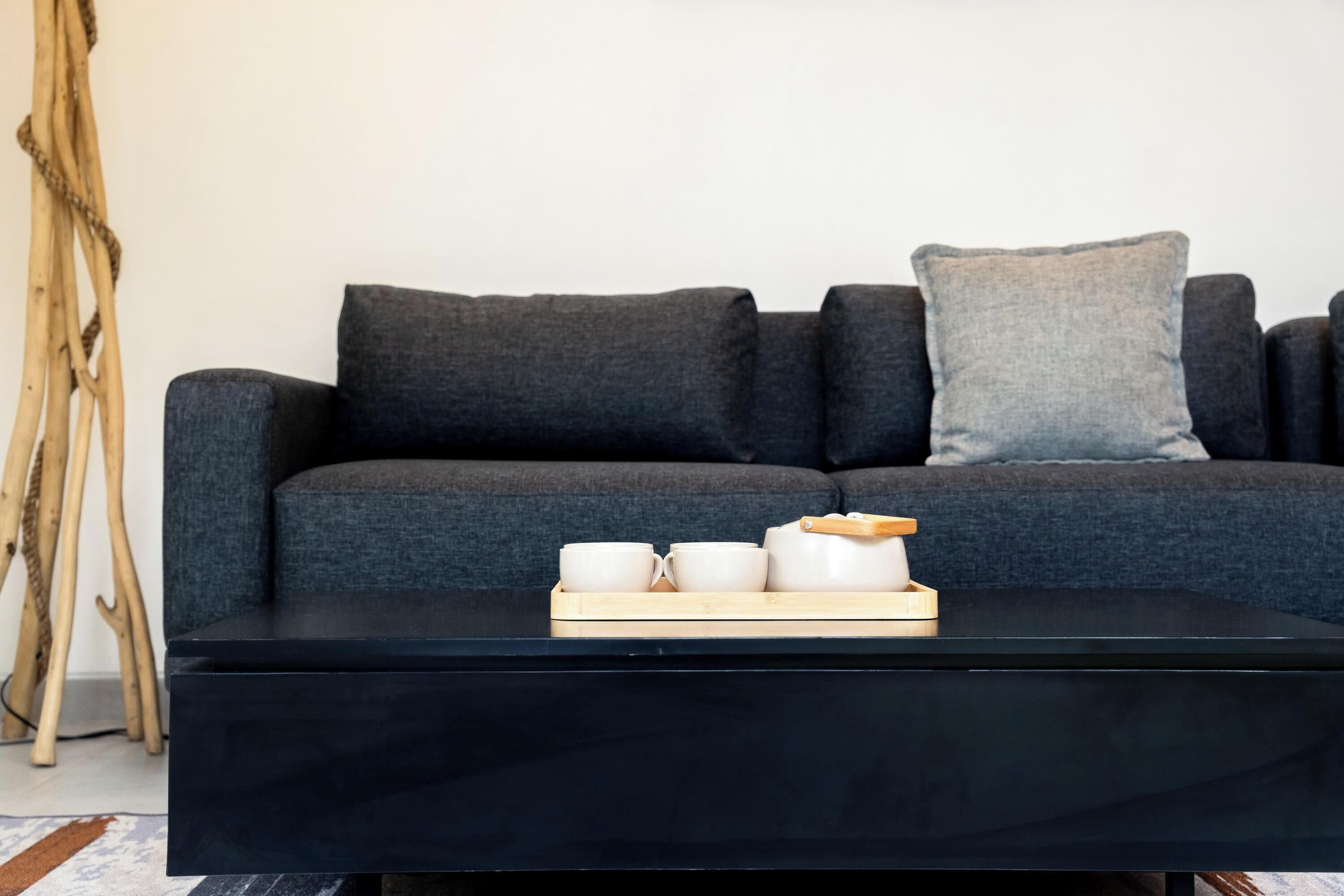 Una sala moderna con un sofá gris oscuro, una mesa de centro negra y una mesita de noche de madera con tazas y un bol.