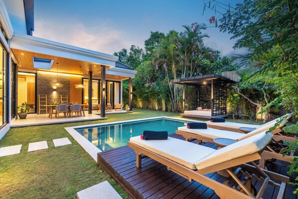 A modern outdoor pool area with lounge chairs, a wooden deck, and a glass-enclosed patio. - Tropical Villa Raya (Ungasan)