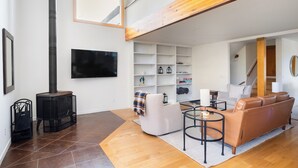 A modern living room with a fireplace, a sofa, a TV, and a staircase leading to an upper level. - Glen Ellen Wine Country Retreat w/pool, hot tub, Bocce, tree-house & more (Glen Ellen)
