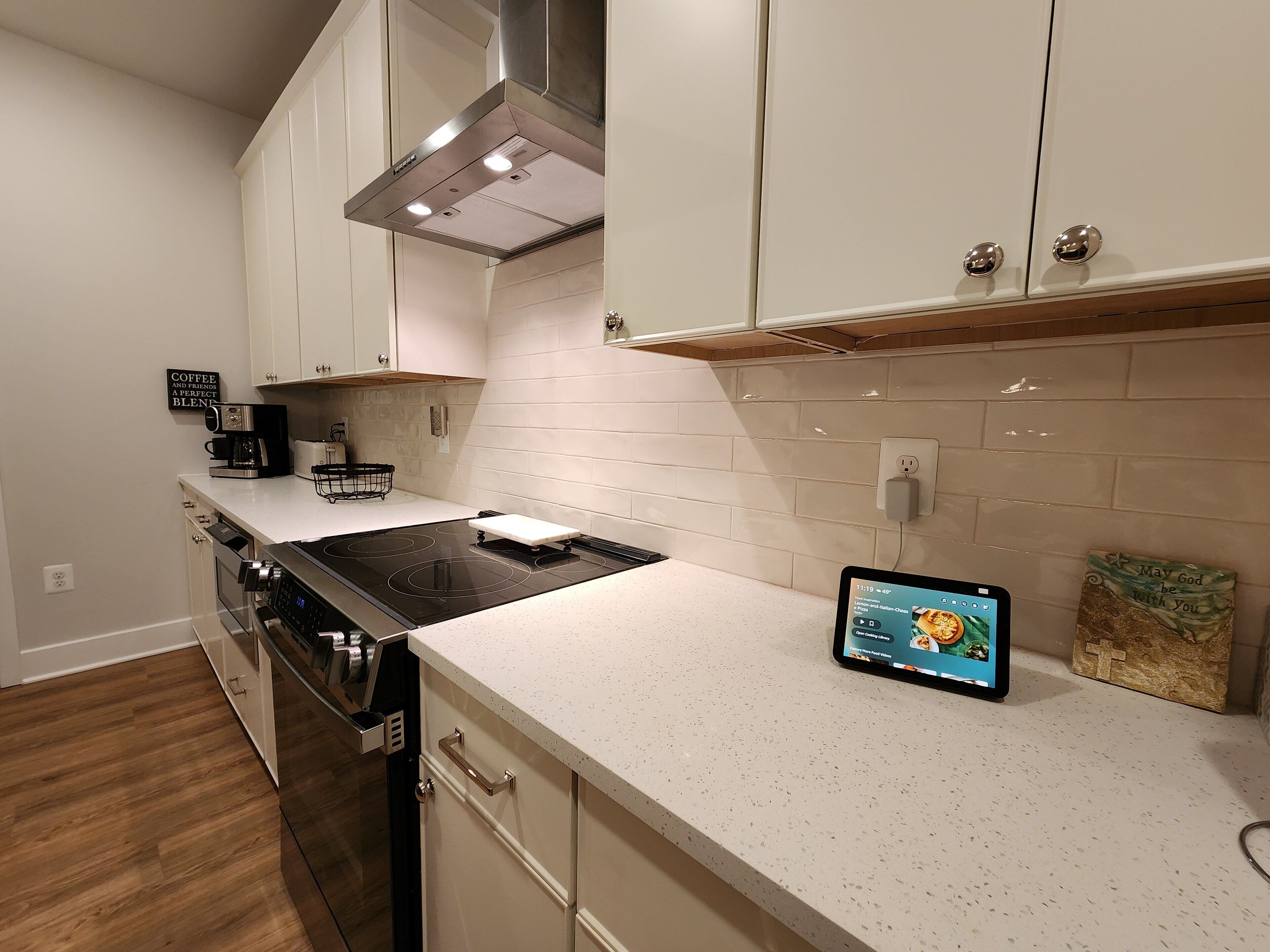 Cocina moderna con gabinetes blancos, campana de acero inoxidable, horno integrado y una tableta en la encimera.