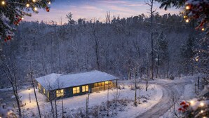 Una cabaña con techo nevado, rodeada de un bosque, accesible mediante un camino de tierra.