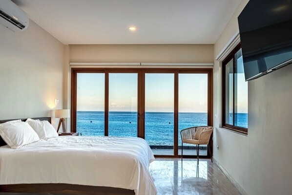 Ein Schlafzimmer mit einem großen Bett, einem Sessel und Meerblick durch eine Schiebetür.