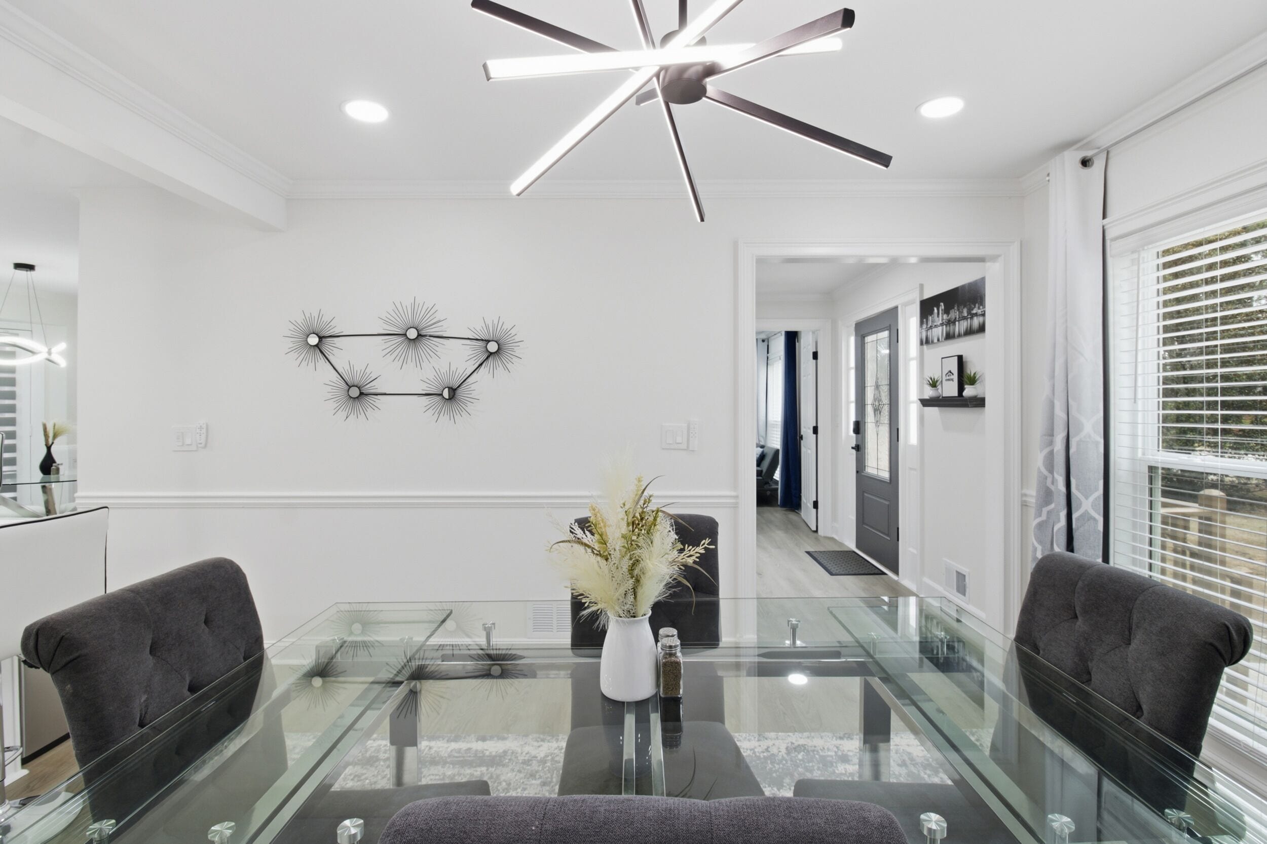A modern dining area with a glass table, two dark chairs, a vase with dried flowers, and a ceiling fan.