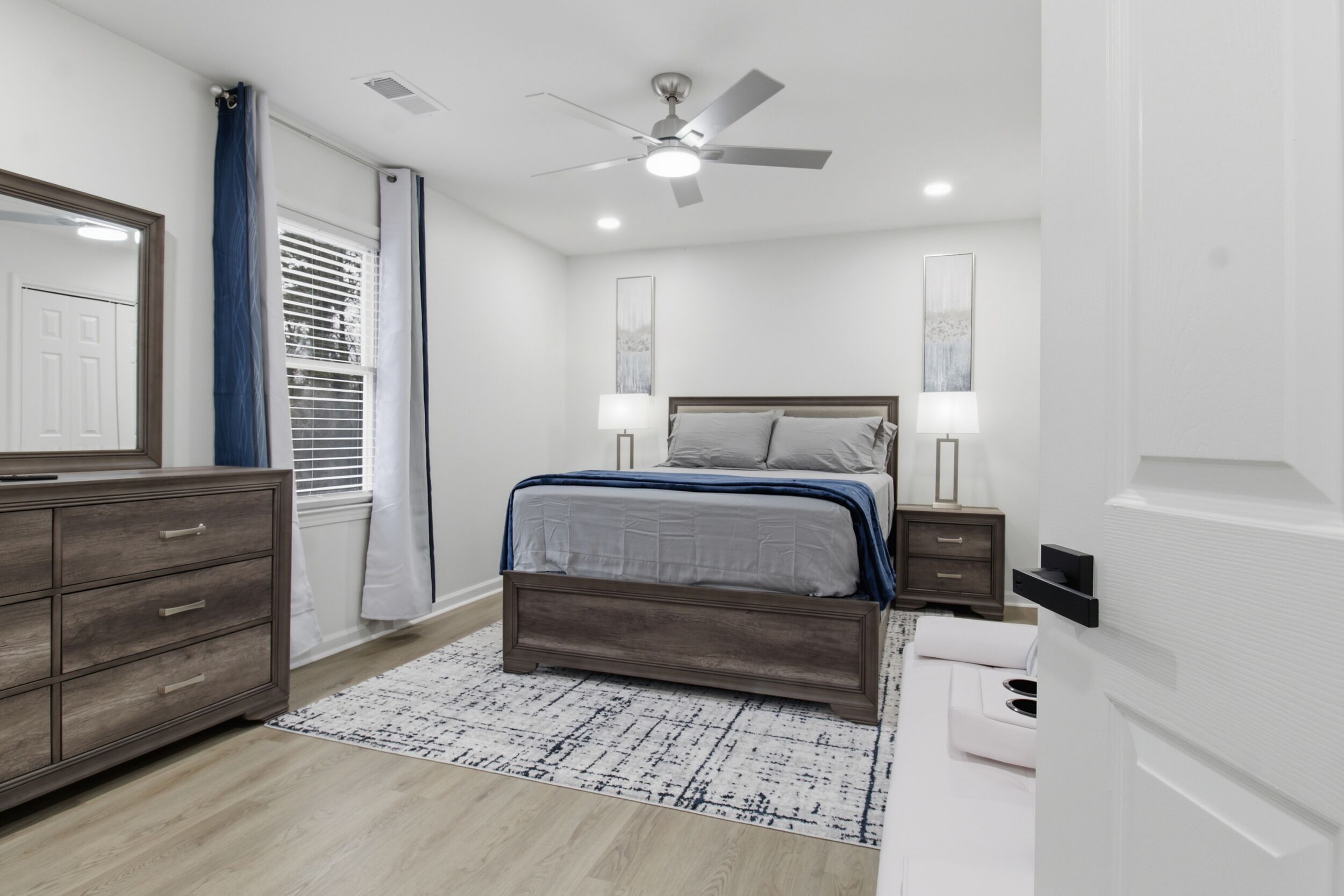 Un dormitorio con cama, mesitas de noche, cómoda, ventilador de techo y ventana con persianas.