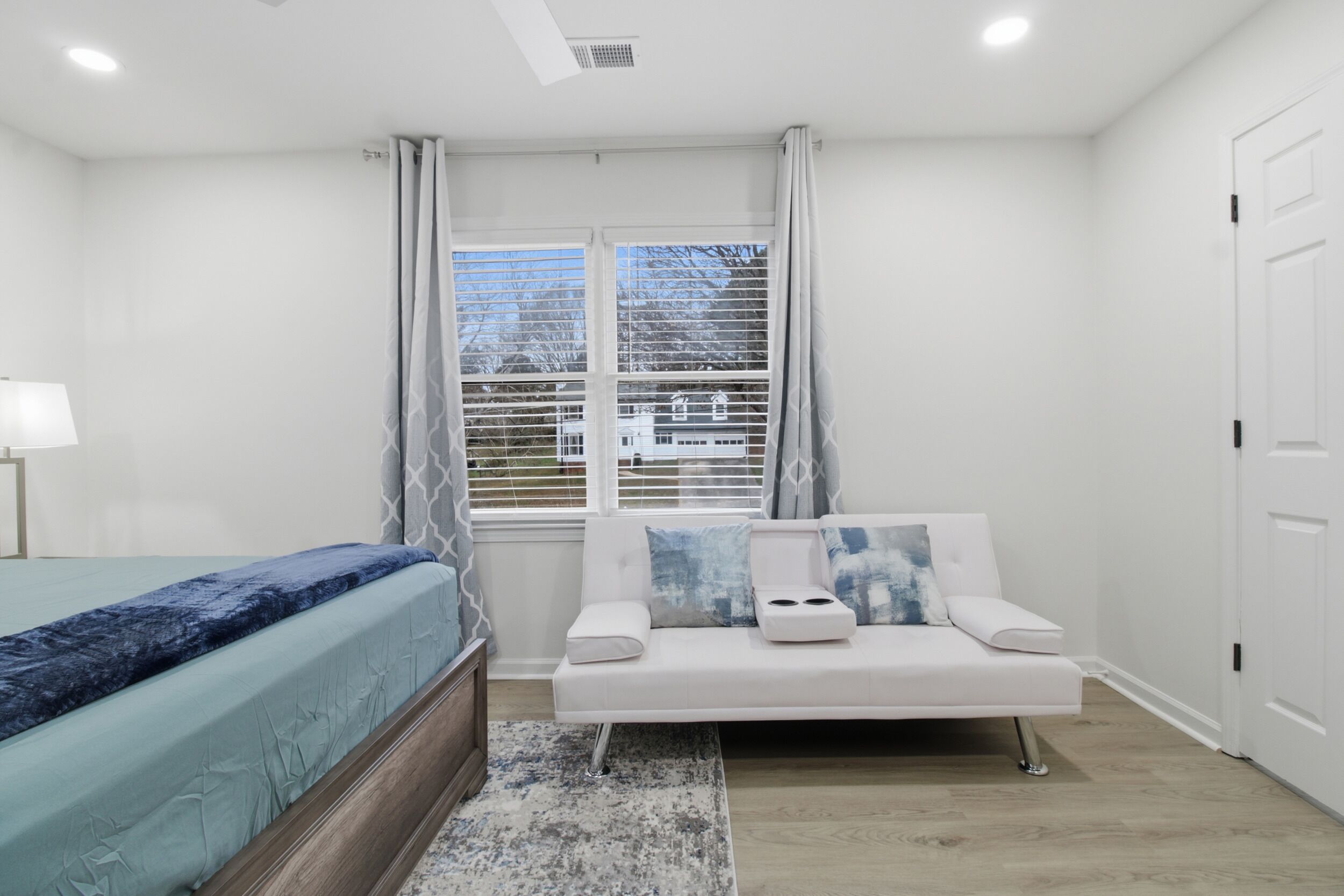 A bedroom with a bed, a white sofa, a window with blinds, and a door.
