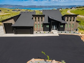 A modern house with a garage, a driveway, and a view of a lake and mountains. - Stunning Lake & Mountain Views! NEW 6BR Home | Hot Tub, Private Beach | Sleeps 26 (Saint Charles)