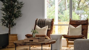 Un salon chaleureux avec une grande fenêtre, un fauteuil en cuir marron, une table basse en osier et une plante en pot.