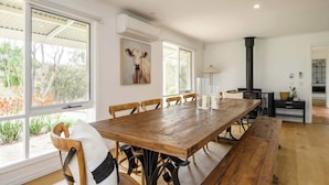 Un vaste espace dédié à la salle à manger, avec une table et des chaises en bois, de grandes fenêtres donnant sur la verdure, un tableau accroché au mur et un poêle noir en arrière-plan.