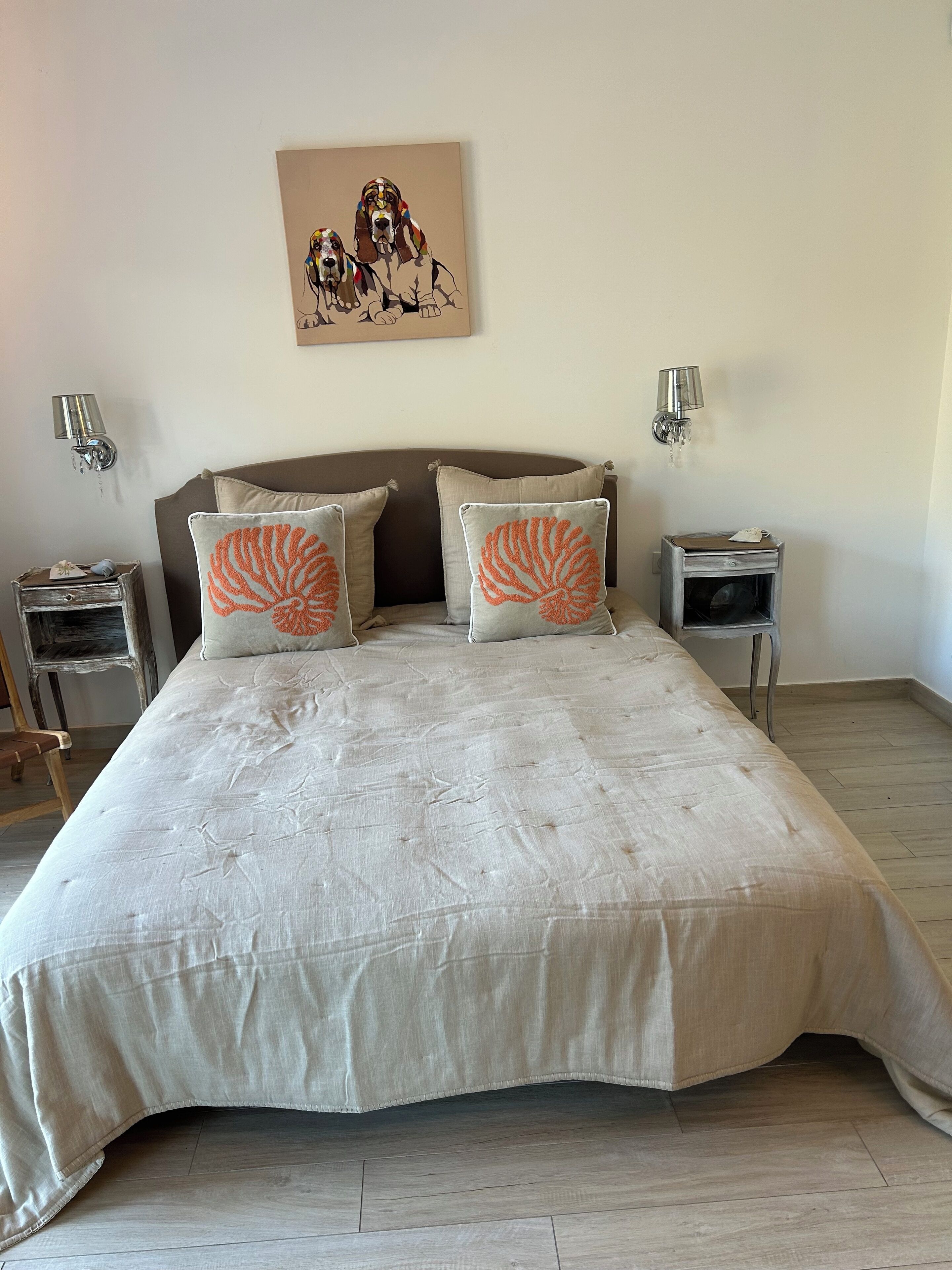 Un letto grande con una coperta beige e due cuscini con motivi a conchiglia arancioni, affiancato da due comodini con lampade e un piccolo tavolino.