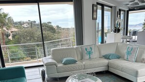 Een moderne woonkamer met een witte modulaire bank, kussens in teal-tint, en een ronde salontafel. Grote ramen bieden uitzicht op een balkon en omliggend groen.
