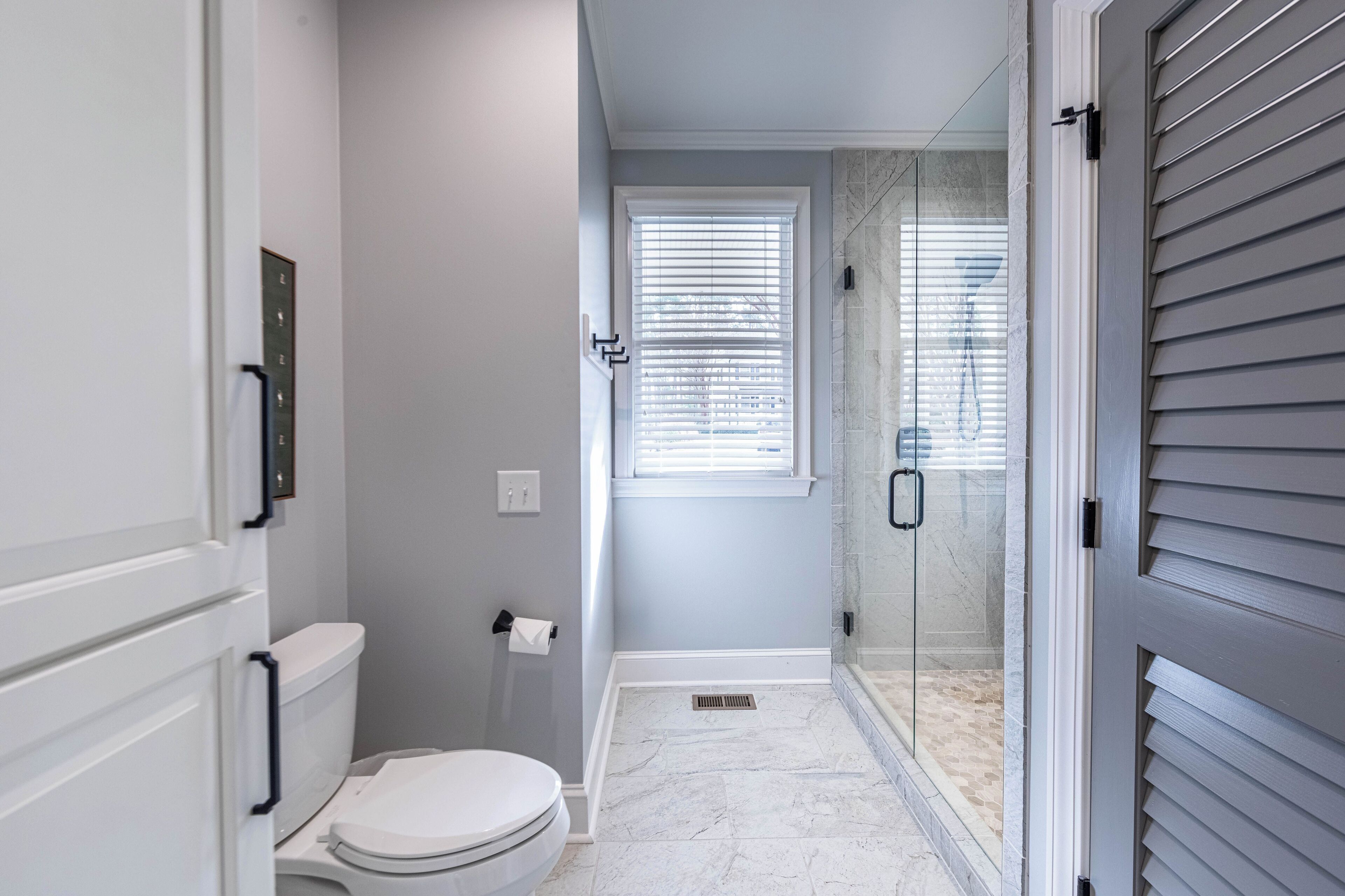 Une salle de bain comprenant des toilettes, une douche fermée par des parois en verre et une fenêtre avec des stores.