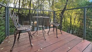 Une terrasse en bois, agrémentée de chaises en métal et d’une petite table, entourée d’une rambarde métallique, et offrant une vue sur une zone boisée.